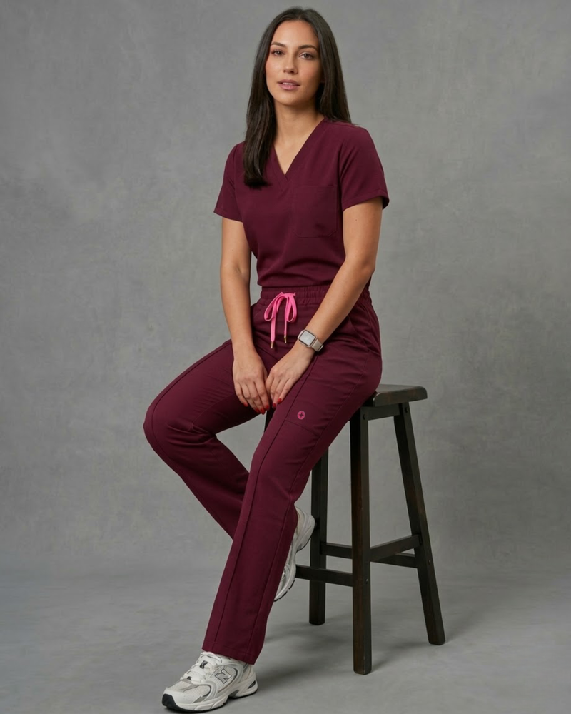 Woman wearing a maroon scrub set sitting on a stool against a gray background